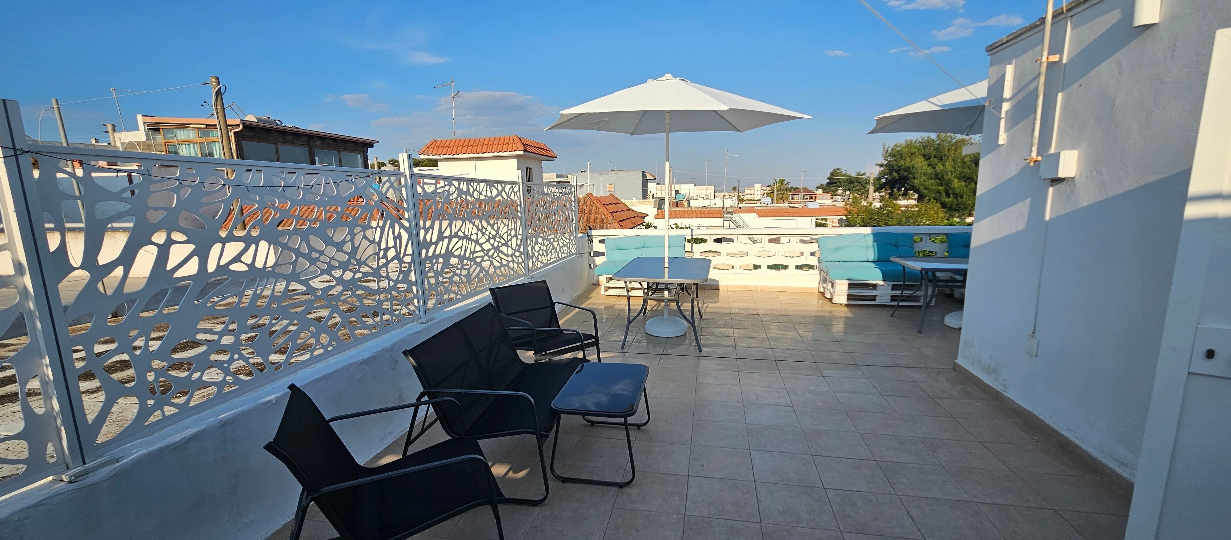 Terrazza condivisa con ringhiera in ferro intagliato bianco, ombrelloni, divani e tavoli per cene all'aperto.
